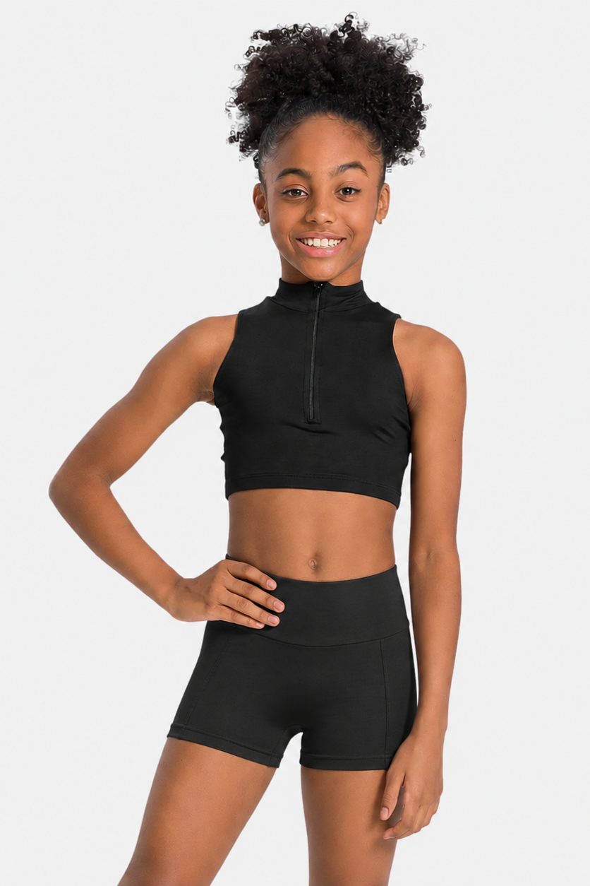 A young girl with curly black hair wearing a black sleeveless mock-neck athletic crop top with a front zipper and matching black high-waisted shorts, posing against a light gray studio background.