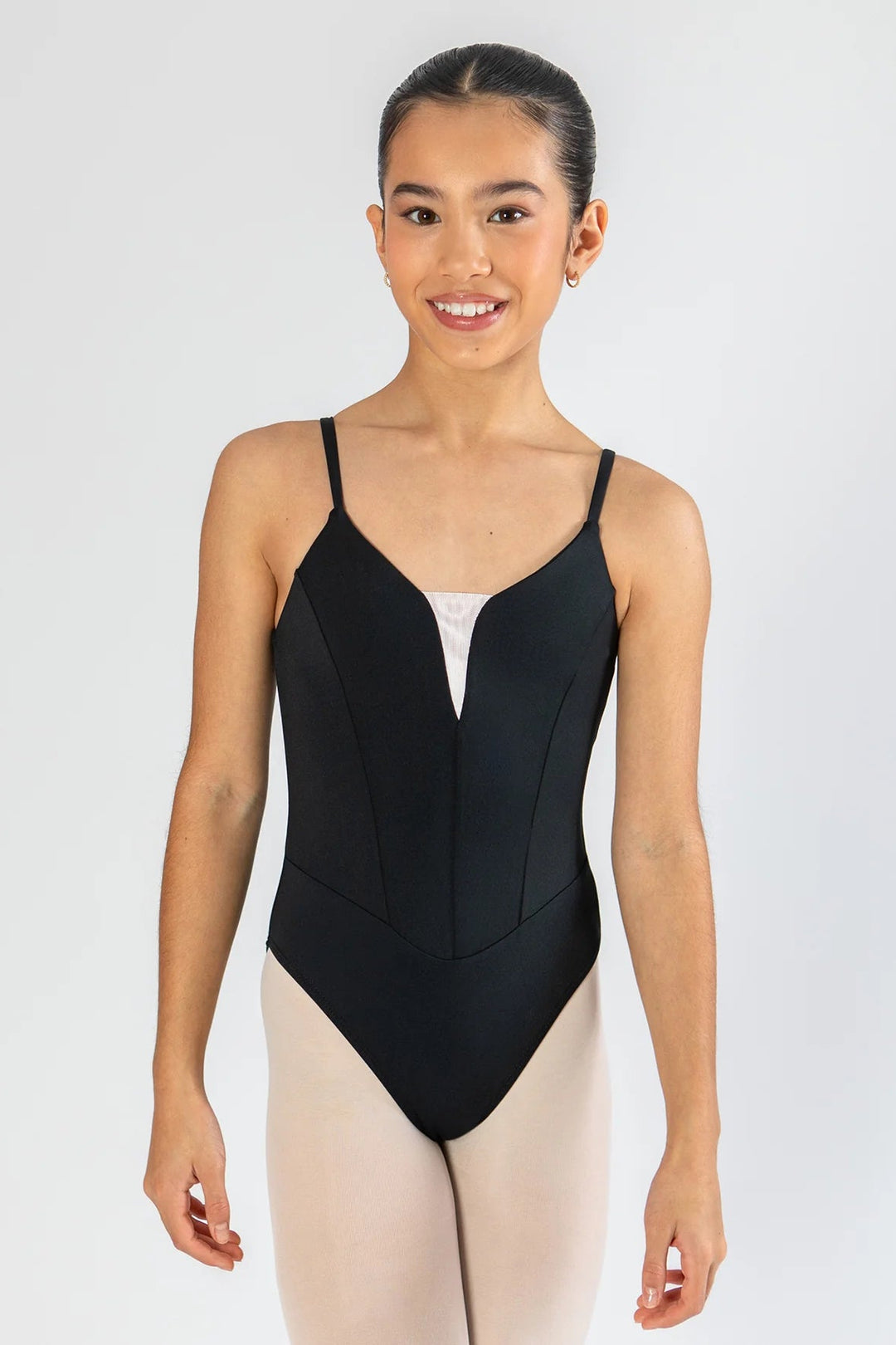 A young female dancer wearing a black bodysuit with thin spaghetti straps and a split neckline, paired with light pink tights, standing against a light gray studio background.