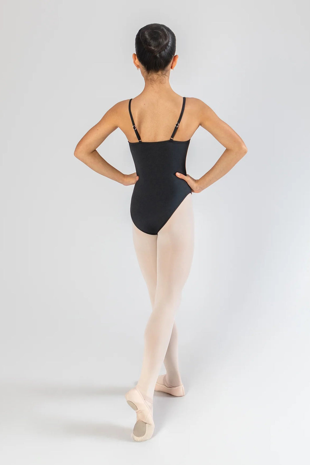 A young ballerina is shown from behind wearing a black leotard, light pink tights, and pointe shoes against a light gray studio background.